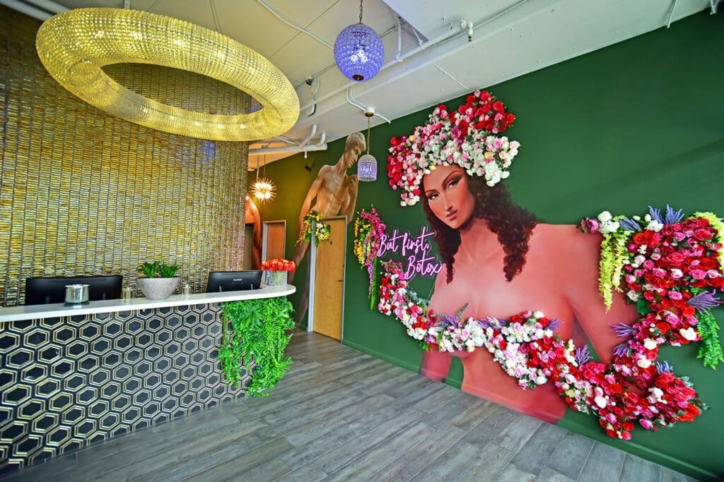 Modern medical spa reception with a green wall, floral goddess mural, neon sign, and gold ring chandelier.